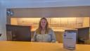 A woman (Riann) behind the counter. She looks straight into the camera. She is seated between two computer screens, and her name card is on the right side of the counter.
